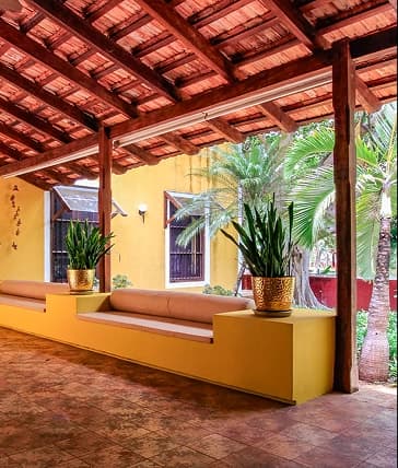 Un pasillo exterior techado con tejas de barro y vigas de madera, flanqueado por paredes amarillas, bancas integradas del mismo color y macetas con plantas largas.